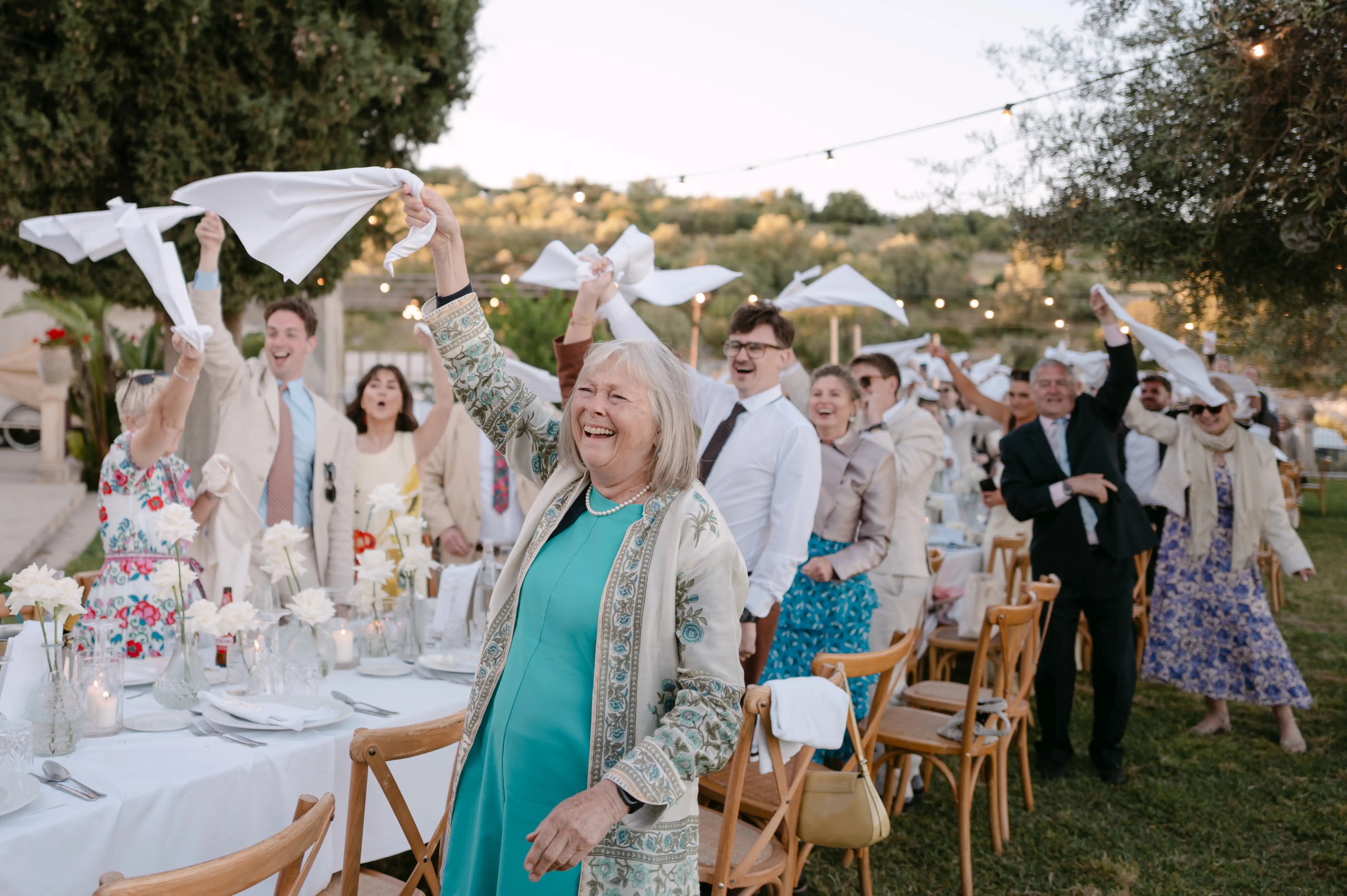 Wedding guests waving white napkins and cheering during an elegant outdoor dinner reception in Sicily.